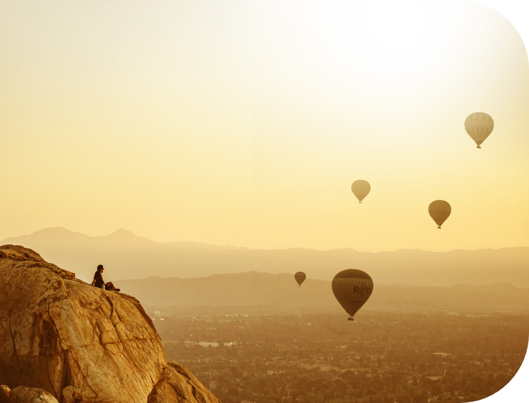 Person sitting on mountain side looking at hot air balloons